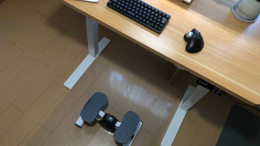 電動昇降デスクを高く設定し、ステッパーを踏みながら作業している足元と、高さ108cmを表示したコントローラー。配線はケーブルトレーで整理されている