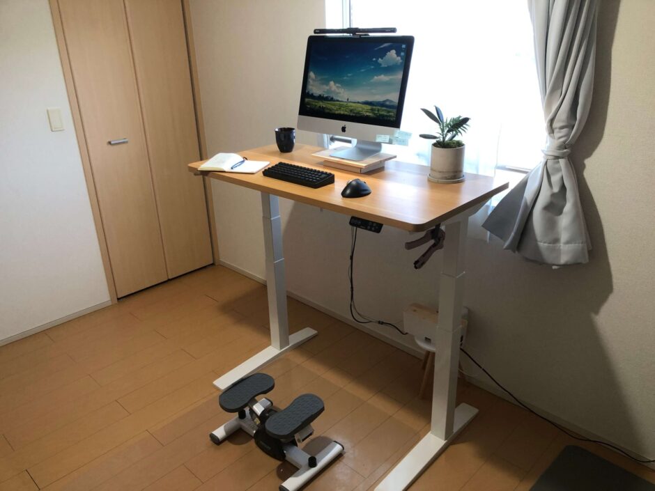 扉から見た6畳の在宅ワーク環境。電動昇降デスクとステッパーを設置し、デスク上にはコーヒー、メモ帳、観葉植物を配置している様子